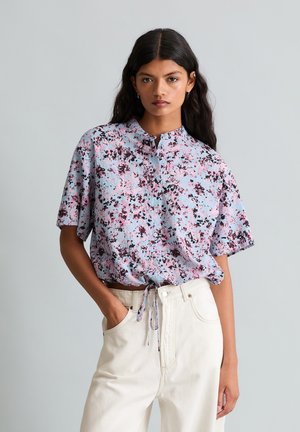 Frau trägt eine kurzärmelige, hellblaue und rosa florale Bluse mit Kordelzug in der Taille, kombiniert mit hoch taillierten, offwhitefarbenen Hosen.