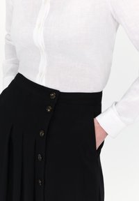 Witte linnen blouse met een knoopsluiting, gecombineerd met zwarte broek met hoge taille met plooien en decoratieve bruine knopen.