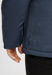 Et nærbillede af en persons hånd, der hviler på kanten af en marineblå jakke, hvor plejeinstruktioner er trykt på stoffet.