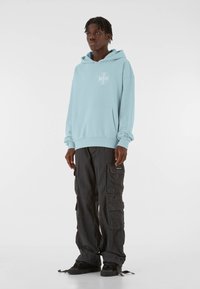 Svetlomodrá mikina s kapucňou a predným vreckom, s bielym grafickým dizajnom, kombinovaná s čiernymi cargo nohavicami s viacerými vreckami a voľným strihom.