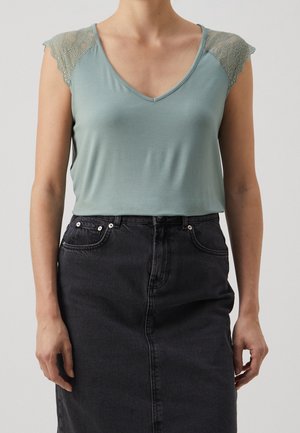 Femme portant un haut sans manches vert clair avec des épaules en dentelle et une jupe en jean noire taille haute, debout sur un fond uni.