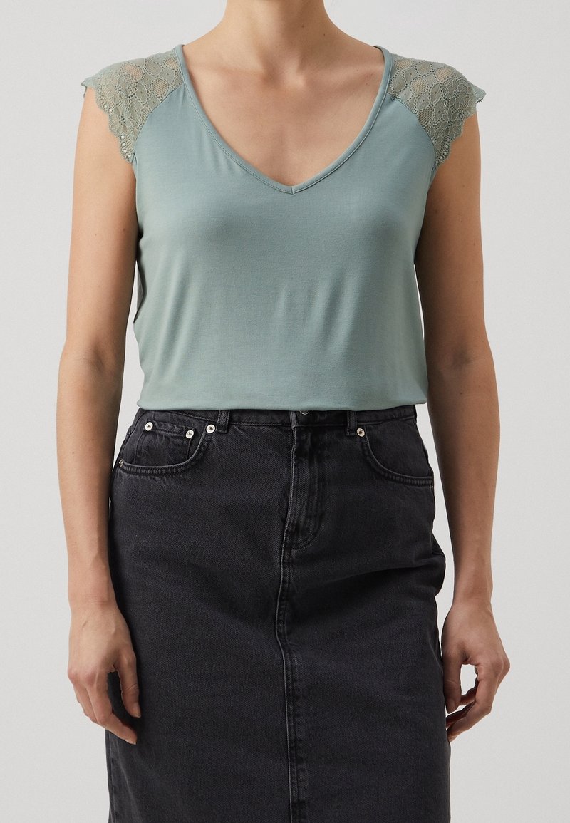 Femme portant un haut sans manches vert clair avec des épaules en dentelle et une jupe en jean noire taille haute, debout sur un fond uni.