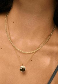 Collier à multiple couches en ton or, présentant un pendentif rond texturé sur une chaîne délicate et une chaîne torsadée, en gros plan sur la peau.