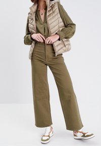 Gilet matelassé beige, blouse vert olive, pantalon taille haute vert olive, et baskets blanches avec des accents verts et marron.