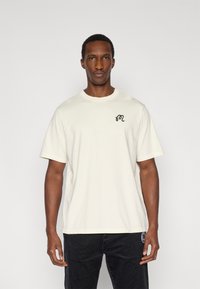 Άσπρο βαμβακερό t-shirt με κοντά μανίκια, στρογγυλή λαιμόκοψη και μαύρο κεντημένο λογότυπο στο στήθος, σε συνδυασμό με μαύρο παντελόνι από κορδόνι.