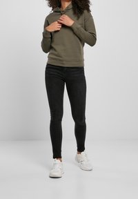 Sweat à capuche vert olive avec capuche à cordon de serrage, associé à un jean skinny noir et des chaussures de sport blanches. Tissu lisse avec une coupe décontractée.
