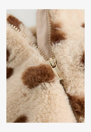 Beige faux karusnahk, millel on pruunid laigud, koos kuldse tõmblukuga ja sileda tekstuuriga. Šotitud lähedalt tõmbluku ja kanga detail.