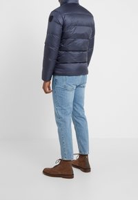 Marinblå puffjacka med hög krage, med horisontell quiltad design. Bärs med ljusblå jeans och bruna mockastövlar.