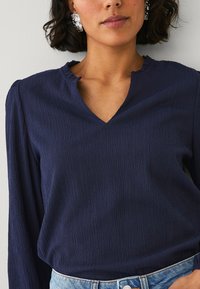 Blusa navy in tessuto texturizzato con un delicato scollo a V, maniche lunghe e spalle arricciate, abbinata a jeans in denim blu a vita alta.