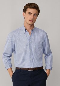 Joven vestido con una camisa de botones a rayas azul claro con las mangas enrolladas, metida en pantalones oscuros con un cinturón marrón, de pie con las manos en los bolsillos.