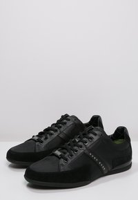 Mustat Hugo Boss -nauhalliset tennarit, joissa on sekoitettu materiaalitoppi, mokka-yksityiskohdat ja brändätty sivuraita, aseteltuna vierekkäin valkoisella pinnalla.