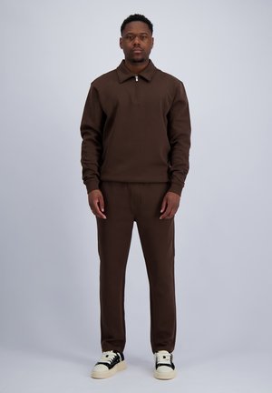 Man staat, draagt een bruin zip-up top met lange mouwen en bijpassende broek met zwart-witte sneakers tegen een effen grijze achtergrond.