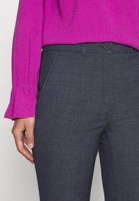 Gros plan d'une personne portant un pantalon ajusté gris foncé et un haut à manches longues violet vif avec des poignets évasés, main détendue le long du corps.