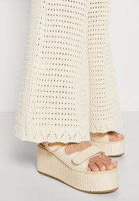 Woven platformowe sandały w kolorze kremowym z szerokim regulowanym paskiem i teksturowaną powierzchnią. Połączone z luźnymi, dzianinowymi, kremowymi spodniami o rozszerzanych nogawkach.