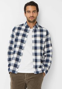 Camicia a quadri blu e bianca con bottoni, realizzata in morbido cotone, maniche lunghe e tasche sul petto. Indossata sopra una t-shirt bianca.