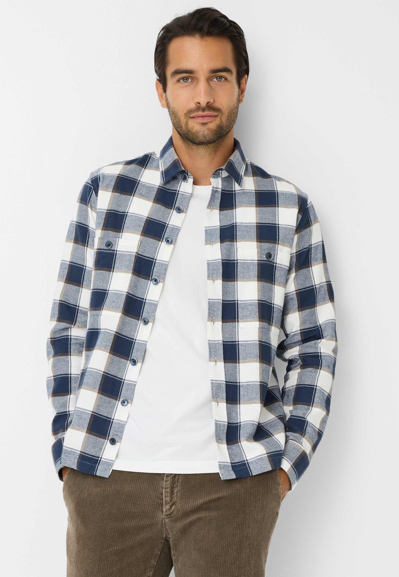 Camicia a quadri blu e bianca con bottoni, realizzata in morbido cotone, maniche lunghe e tasche sul petto. Indossata sopra una t-shirt bianca.