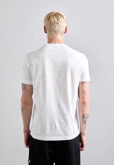 Blond-haired person with tattoos on both arms wearing a plain white t-shirt and black pants, facing a light gray wall.