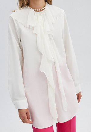 Biała bluzka z długim rękawem z warstwowymi falbanami z przodu i perłowym naszyjnikiem, w połączeniu z jasnoróżowymi spodniami, na osobie stojącej na białym tle.