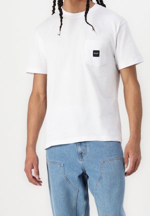 Vit bomullst-shirt med rund hals, korta ärmar och en vänster bröstficka med en svart etikett med texten "HUF".