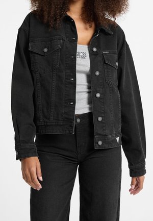 Person wearing a black denim jacket over a gray Guess Jeans tank top and black high-waisted pants, standing against a white background.