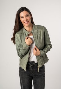 Giacca sintetica verde chiaro con zip frontale, scollo rotondo, accenti a coste sulle spalle e tasche laterali, abbinata a jeans neri.