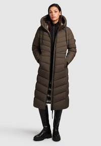 Garšs olīvzaļš pūkains mētelis ar kapuci, ar rāvējslēdzēju un diognālā šuvuma rakstu. Iezīmējas ar melniem akcentiem un tiek kombinēts ar līdz ceļiem melnām zābakiem.