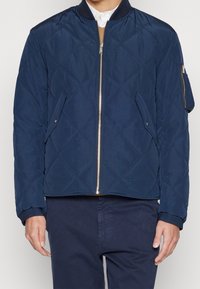 Námořnicky modrá prošívaná bombera s zapínáním na zip, žebrovaným límcem a manžetami, dvěma bočními kapsami a kapsou na zip na rukávu.