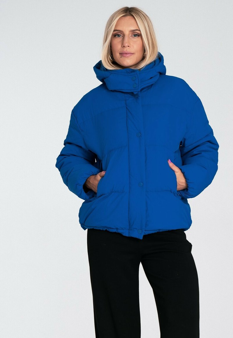 Niebieska kurtka puchowa z wysokim kołnierzem, zapięciem na guziki i bocznymi kieszeniami. Posiada pikowany wzór oraz luźny, oversize'owy krój.