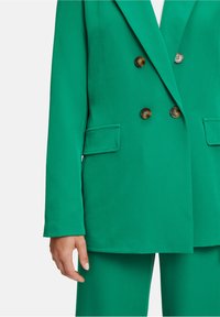 Mujer con blazer verde brillante de doble botonadura con botones de carey y pantalones a juego, con la mano relajada al lado.