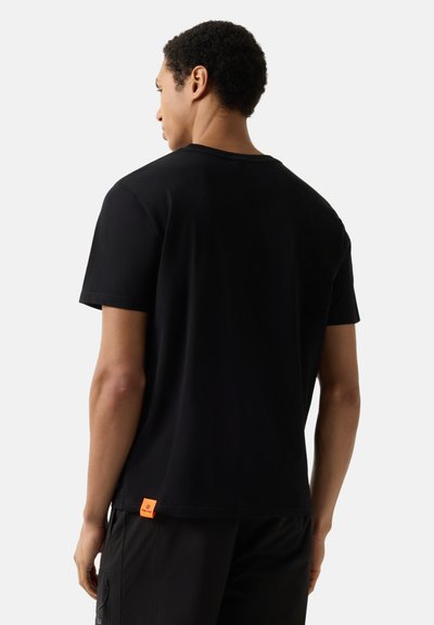 Homme aux cheveux courts et bouclés portant un t-shirt noir uni avec une étiquette orange sur le bas du dos, vu de dos sur un fond blanc.