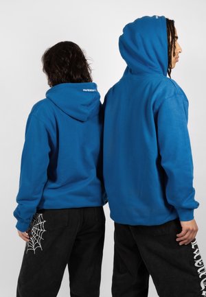 Twee mensen staan rug aan rug, gekleed in blauwe hoodies en zwarte broeken, de een met een spinnenwebontwerp op de zak, de ander met witte tekst op het been.