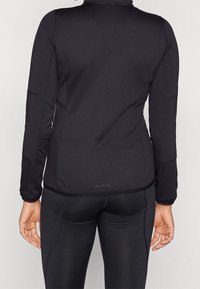 Chaqueta deportiva negra con mangas largas, hecha de un material suave. Presenta paneles texturizados y un pequeño logotipo en la parte baja de la espalda.
