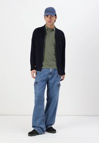Cardigan navy cu fermoar, polo măsliniu, blugi cargo lejeri de culoare albastru deschis, adidași negri și șapcă albastră. Design simplu, cu accente minimale.