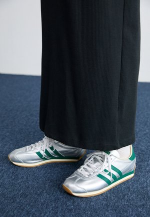 Person iført hvide og grønne sneakers med hvide sokker og en lang sort nederdel stående på blåt tæppe.