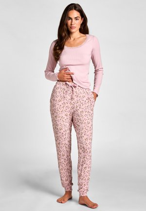 Vrouw in een roze top met lange mouwen en roze bloemen-prints pyjamabroek met koord, blootsvoets staand tegen een effen grijze achtergrond.