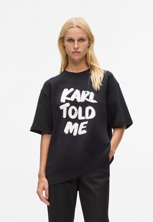 Black oversized t-shirt with short sleeves featuring bold white lettering "KARL TOLD ME." Made of cotton, smooth texture, relaxed fit.