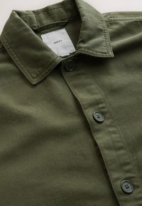 Chemise verte boutonnée avec col, comportant une étiquette blanche avec le texte « NEXT » à l’intérieur du col.