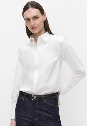 Frau mit glatten braunen Haaren, die ein weißes Hemd mit Knöpfen trägt, das in dunkelblaue Jeans mit einem schwarzen Gürtel gesteckt ist.