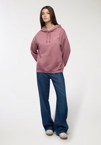 Felpa cappuccio rosa con tasca frontale, abbinata a jeans blu a gamba larga. La felpa presenta un piccolo logo sul petto.