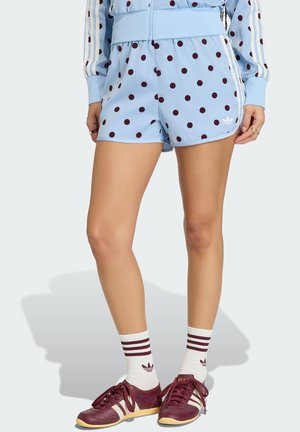 Benen met lichtblauwe korte broek met witte stippen, mouwen van een bijpassend jasje, witte sokken met bordeauxrode strepen en bordeauxrode Adidas-sneakers.