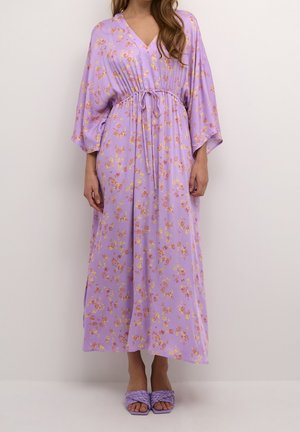 Femme portant une longue robe fleurie lavande avec une taille à cordon et des manches larges, associée à des sandales tissées à talons assorties.