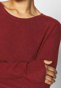 Pull rouge à manches longues avec un col bateau, un tricot texturé et une coupe légèrement ample. Dispose de manches surdimensionnées avec des détails de poignet.