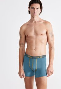 Boxers para hombres en tejido teal con un ajuste ceñido, con costuras amarillas y una cinturilla gris oscuro etiquetada "sloggi." Textura suave, sin patrones.