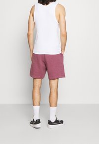 Muž oblečený v bílém tílku, vínových šortkách, bílých sportovních ponožkách a černých teniskách, stojí s rukama v kapsách čelem ke bílé zdi.