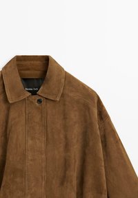 Veste en daim marron avec col et un seul bouton noir visible, partiellement montrée sur un fond blanc.
