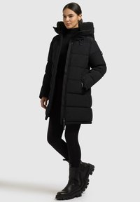 Cappotto imbottito nero con colletto alto e chiusura a zip, dotato di cappuccio testurizzato, tasche laterali e taglio dritto. Abbinato a leggings neri e stivali chunky.