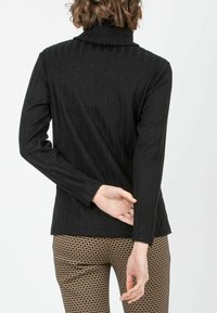 Schwarzer gerippter Pullover mit hohem Kragen, langen Ärmeln und tailliertem Design. Getragen mit gemusterten braunen Hosen, die ein geometrisches Design aufweisen.