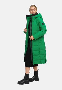 Abrigo acolchado verde con capucha, diseño acolchado, cremallera frontal y dos cremalleras laterales. Combinado con un vestido negro de canalé y botas negras hasta el tobillo.
