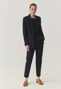 Námořnický blazer se strukturovanými rameny, spárovaný s odpovídajícími zúženými kalhotami s elastickým pasem. Doplněno hnědými semišovými mokasíny.