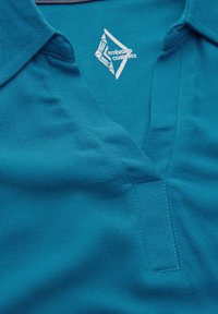 Türazi Polo-Shirt mit V-Ausschnitt, weichem Material, mehrfädig genähtem Kragen und einem weißen Logo auf der Vorderseite.